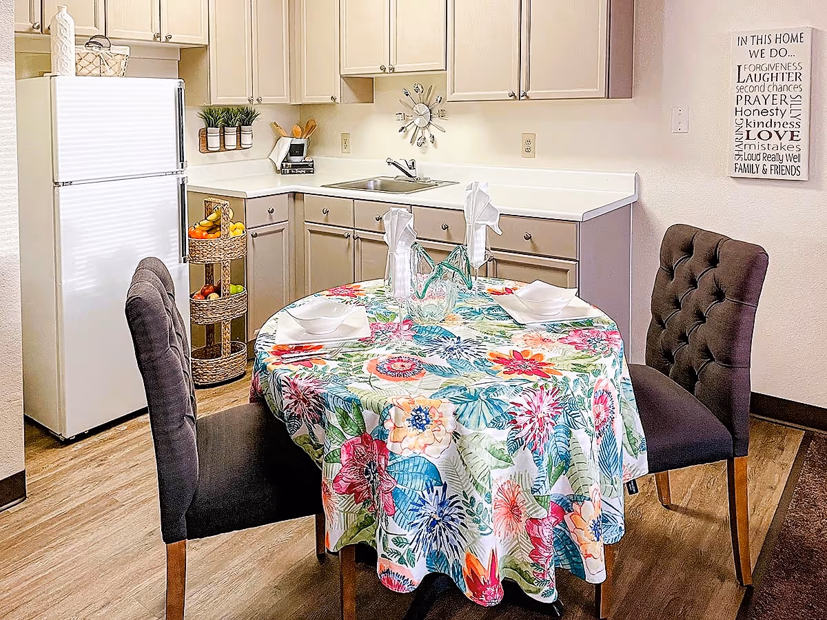 Bright kitchenette with a round table draped in a colorful floral tablecloth, two tufted chairs, cabinets, sink, and a refrigerator.