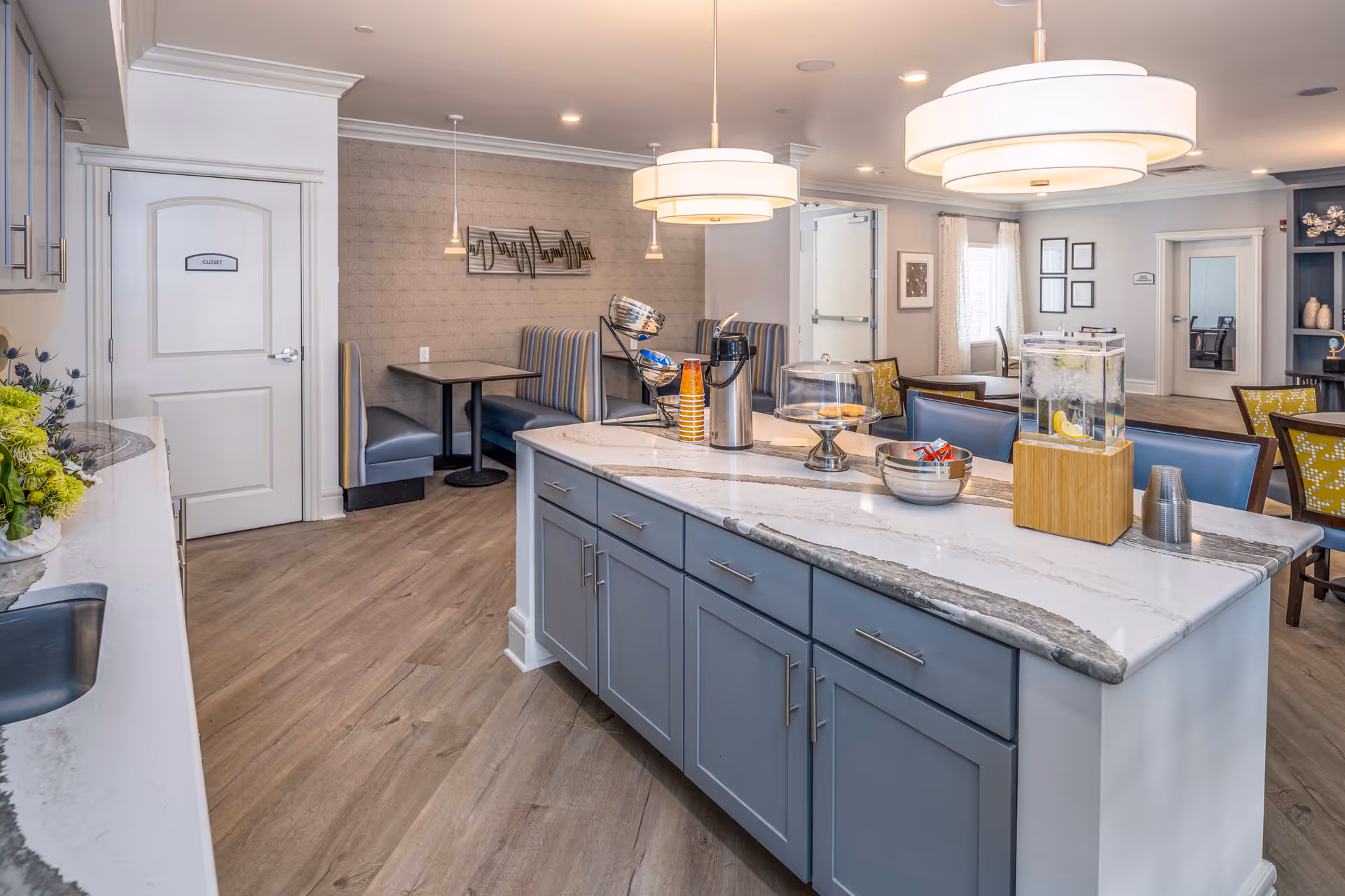 Interior view of a senior living facility dining area with a large kitchen island in the foreground featuring a water dispenser with lemon slices, coffee urn, cups, and snacks. The background shows booth seating, tables with chairs, pendant lights, and a door labeled 'Closet'. The space is well-lit with modern decor and wood flooring.