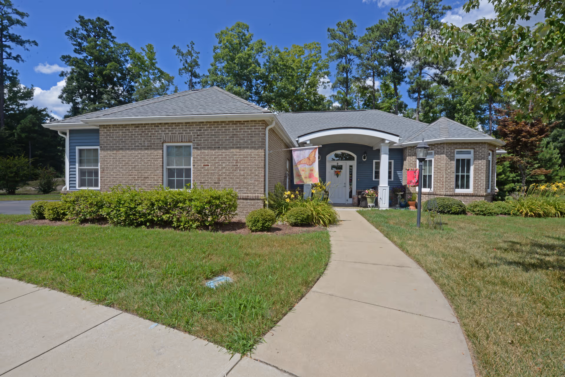 Front exterior view of a single-story brick and siding building with a gray shingled roof, a concrete walkway leading to a white front door under a covered porch, surrounded by green grass, bushes, and trees under a blue sky with some clouds.