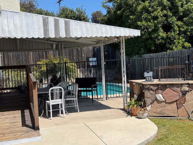 Outdoor patio area with a covered seating space featuring a table and chairs next to a fenced swimming pool. There is a stone structure on the right side with potted plants around it, and trees and a wooden fence in the background.