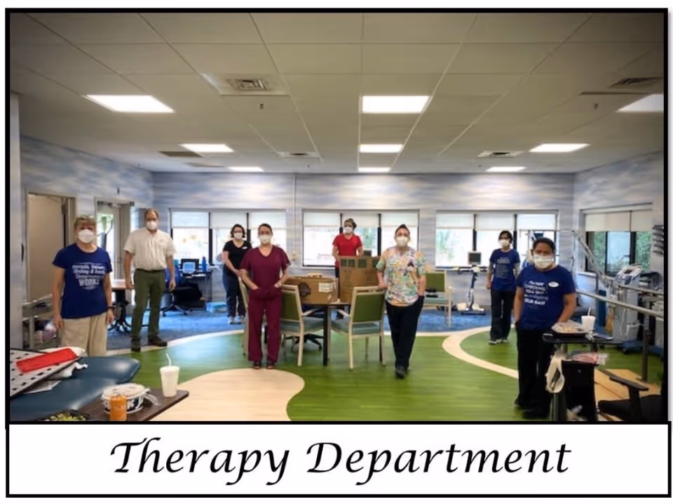 A group of eight healthcare workers wearing masks standing spaced apart in a therapy room with exercise equipment and chairs. The room has large windows, a green and beige floor, and light blue walls. The text below the image reads 'Therapy Department'.