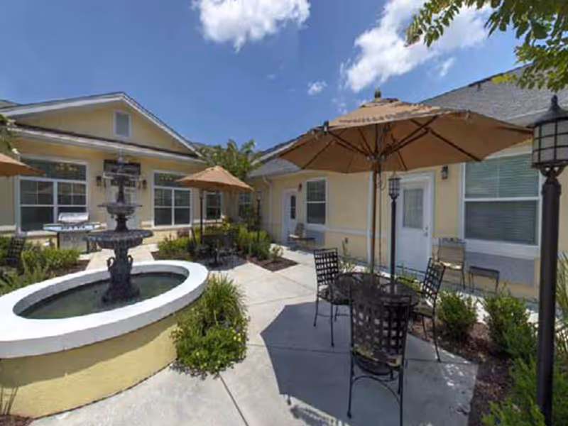 Outdoor courtyard area at Addington Place of Brunswick featuring a circular water fountain, patio tables with umbrellas, chairs, and surrounding greenery under a partly cloudy sky.