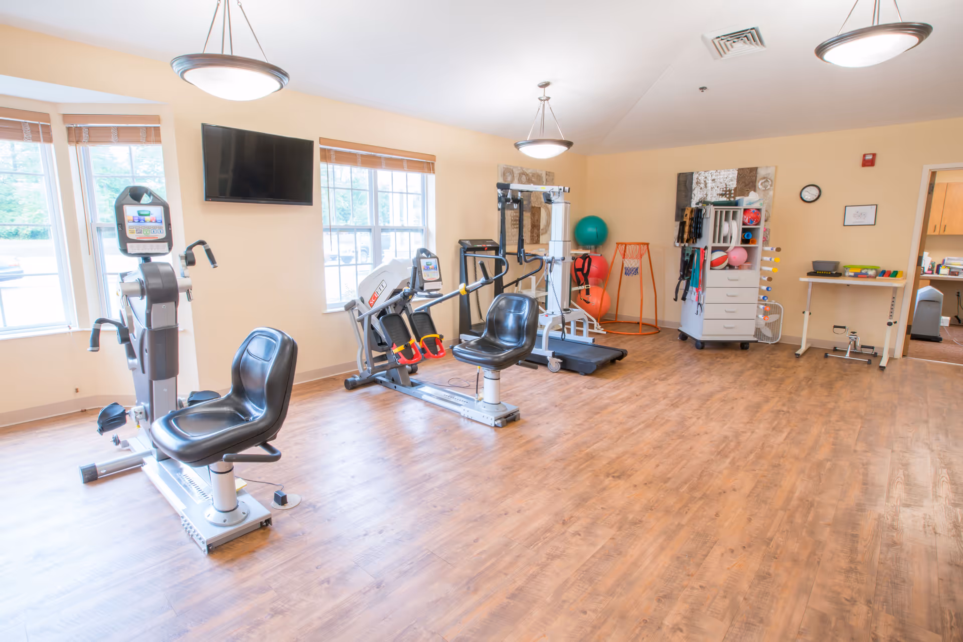 Bright exercise room with seated cardio machines, strength equipment, therapy balls, and storage cabinets.