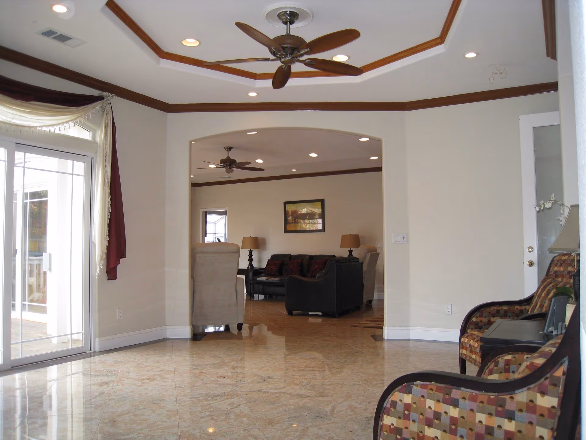 Interior view of a senior living facility showing a spacious room with polished tile floors, a ceiling fan, and recessed lighting. There is a large sliding glass door with curtains on the left, leading to an outdoor area. Through an arched opening, a living room area is visible with armchairs, a sofa, two table lamps, and a framed picture on the wall. Two patterned chairs and a small table with a lamp are on the right side of the foreground.