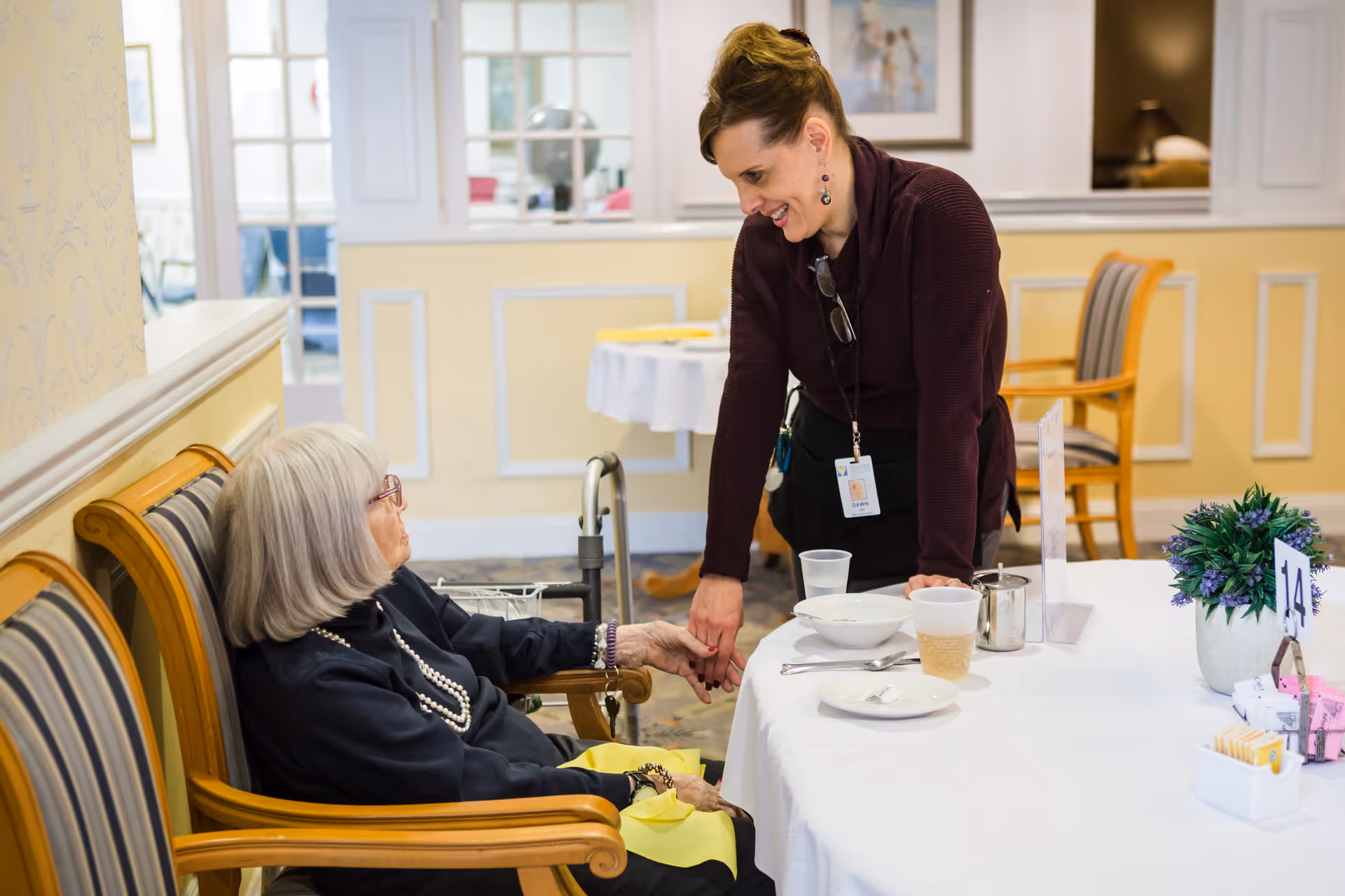 An elderly woman with gray hair and glasses sitting on a wooden bench with striped cushions, holding hands and smiling with a caregiver who is leaning over a table in a warmly lit room. The table has a white tablecloth, dishes, cups, and a small potted plant with a table number 14. The background shows a softly decorated interior with chairs and framed pictures on the wall.