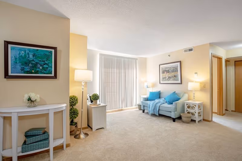 A bright and cozy living room with beige walls and carpet. There is a light blue sofa with blue cushions and a matching throw blanket. Next to the sofa is a white side table with a lamp and a small basket on the floor. A framed picture hangs on the wall above the sofa. On the left side of the room, there is a white console table with decorative boxes underneath and a vase with white flowers on top. A floor lamp and two small potted plants are placed near the console table. Vertical blinds cover a large window letting in natural light.