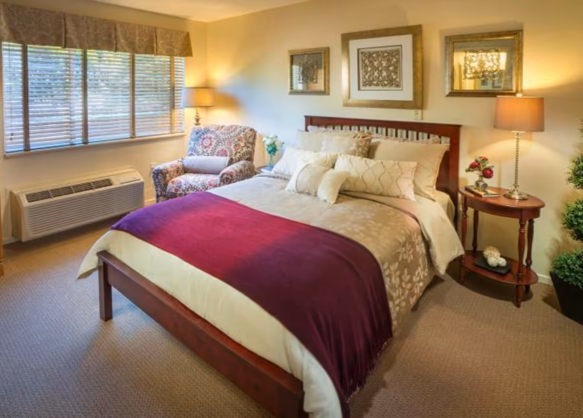 A cozy bedroom with a wooden bed frame, beige and cream bedding, and a purple throw blanket. There is a patterned armchair next to a window with blinds and a valance. Two framed artworks hang above the bed. On the right side, there is a wooden nightstand with a lamp, flowers, and decorative items. The room has beige carpet and soft lighting.