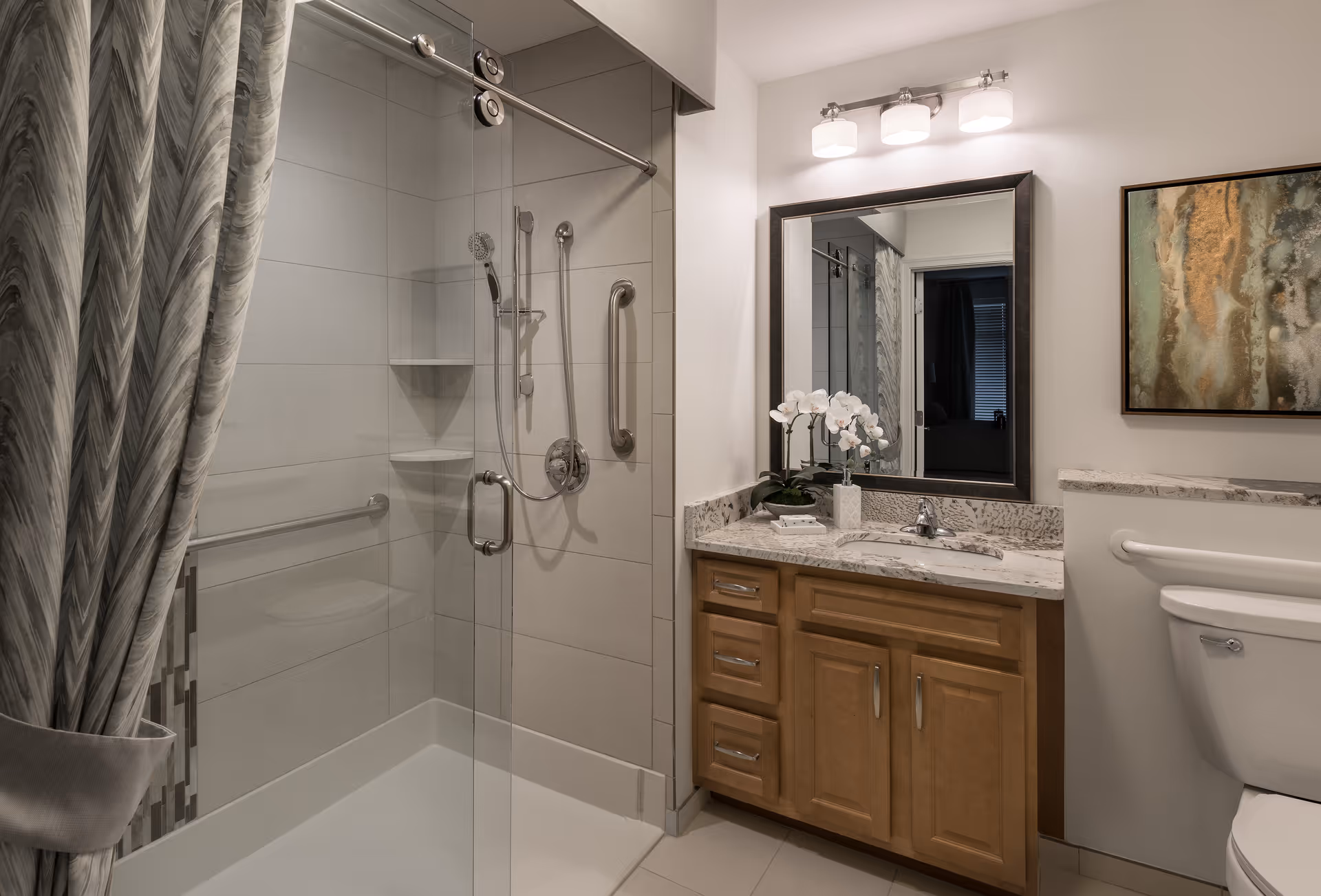 A modern bathroom featuring a glass-enclosed shower with a sliding door, grab bars, and a handheld showerhead. Next to the shower is a wooden vanity with a marble countertop, a sink, a mirror with three light fixtures above it, and a white orchid plant. A toilet is partially visible to the right, and a framed abstract artwork hangs on the wall above it.