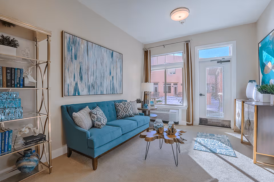 A bright and cozy living room with a teal sofa adorned with patterned pillows, a wooden coffee table with decorative items, a large abstract painting on the wall, a tall metal bookshelf with books and decor, a window with beige curtains, and a glass door letting in natural light.