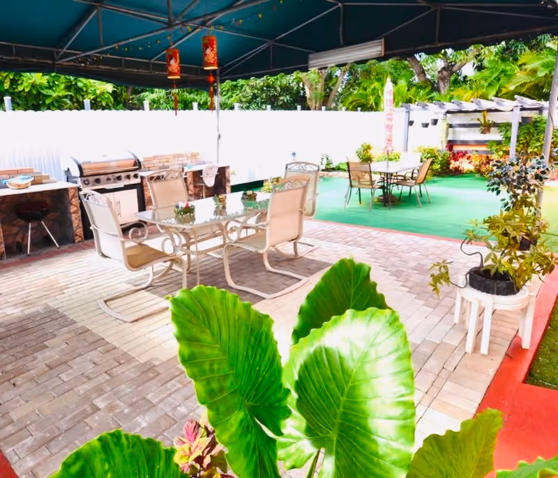 Outdoor patio area with a covered seating space featuring a glass-top table and six chairs. There is a built-in barbecue grill and countertop along the left side. In the background, another table with chairs is set on a green artificial turf area surrounded by plants and trees. Large green leaves are visible in the foreground.