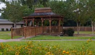 Wooden gazebo sits on a grassy lawn with trees and a building in background.