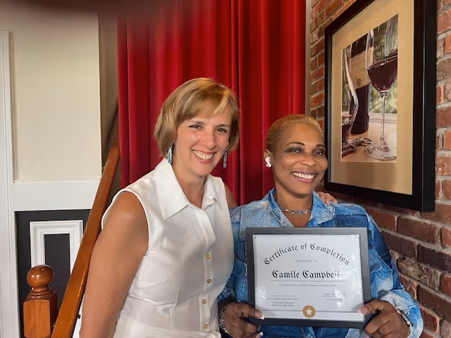Two women standing indoors in front of a staircase and a brick wall with a framed picture of wine glasses. One woman is wearing a white sleeveless blouse and smiling, while the other woman is wearing a blue patterned shirt and holding a certificate of completion with the name Camile Campbell.