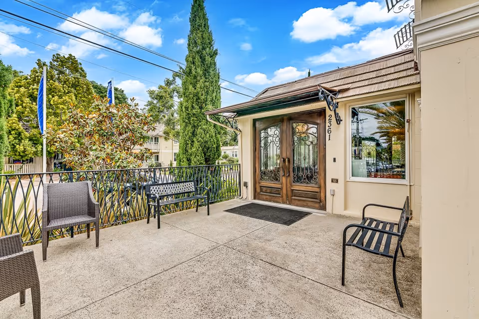 Outdoor patio area with several chairs and benches near the entrance of a building with double wooden doors and decorative ironwork. The building number 23601 is visible next to the door. Trees and a clear blue sky with some clouds are in the background.