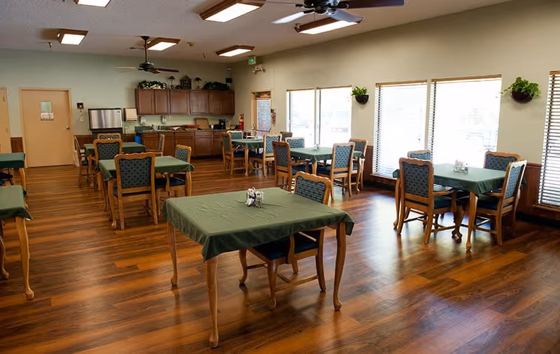 A dining room with several wooden tables covered with green tablecloths and matching wooden chairs with patterned cushions. The room has large windows with blinds allowing natural light to fill the space. There are ceiling fans and overhead lights, and a counter area with cabinets and a microwave in the background.