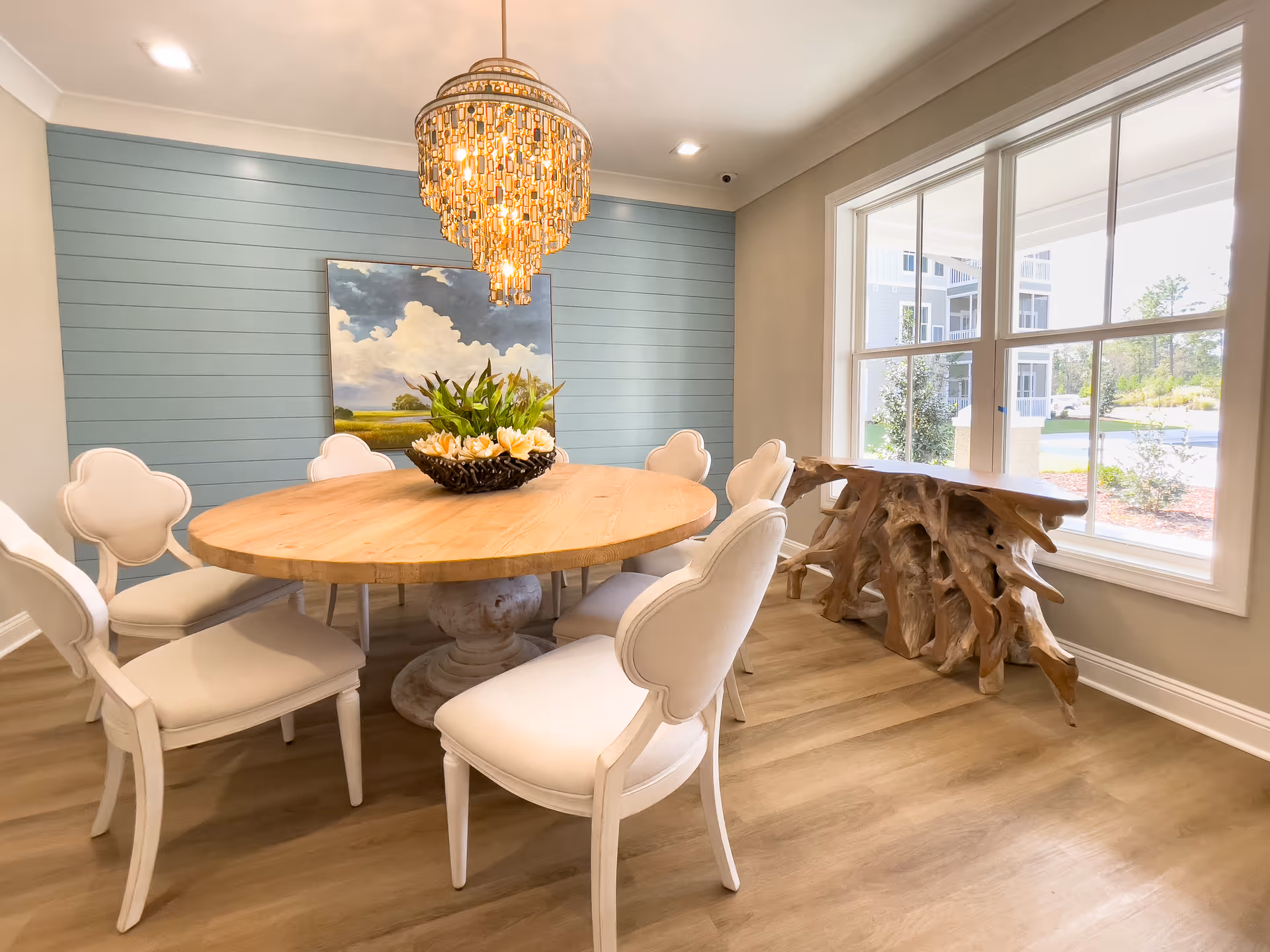 A bright dining room with a round wooden table surrounded by six white upholstered chairs. A decorative bowl with flowers is placed on the table. A large window lets in natural light, and a unique wooden console table with a natural root design sits beneath it. The back wall is painted light blue with horizontal paneling and features a painting of a cloudy sky over a green landscape. A chandelier with multiple hanging crystals illuminates the room.