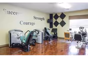 A fitness room with exercise machines including a recumbent bike and other workout equipment. The walls are decorated with motivational words such as 'Success', 'Strength', and 'Courage'. There is a window with a blind and a small table with chairs near it. The floor is wooden.