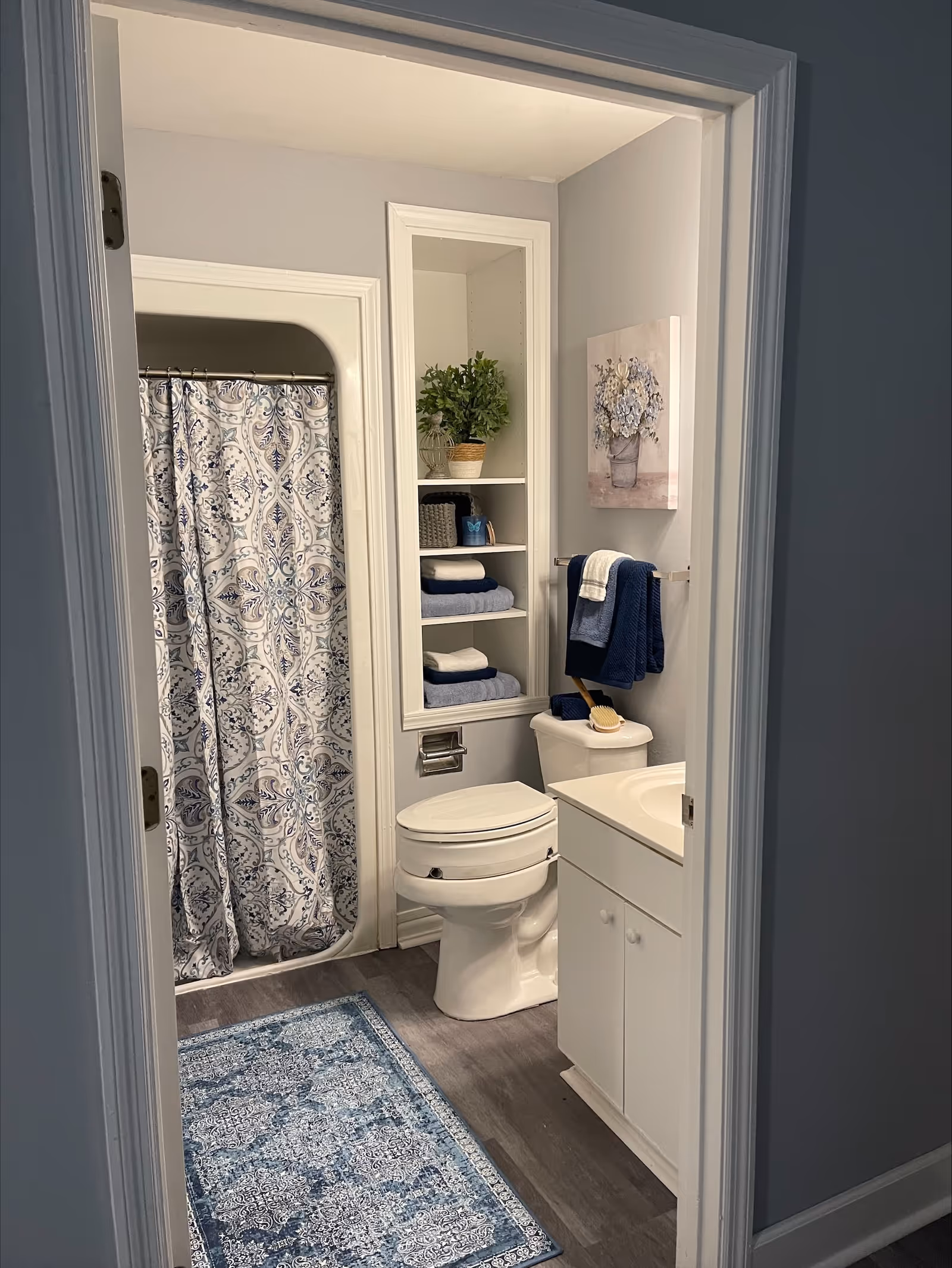 View into a bathroom with a white toilet, white vanity with sink, and a shower with a patterned blue and white curtain. There is a blue and white rug on the floor, built-in shelves with folded towels and a potted plant, a towel rack with blue and white towels, and a floral painting on the wall.