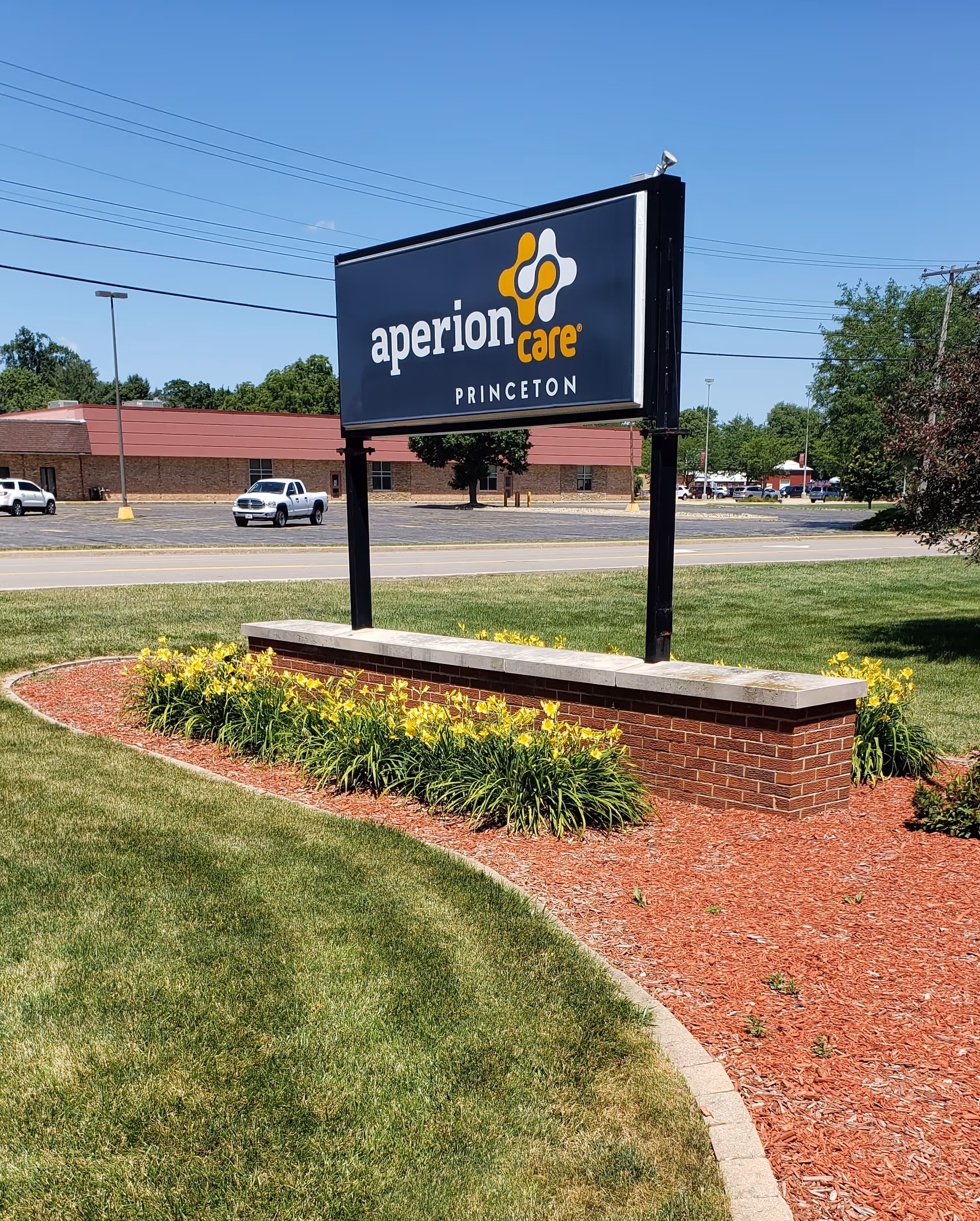 Outdoor sign reading "aperion care PRINCETON" mounted on a brick base with landscaped grass and yellow flowers in front of the facility.