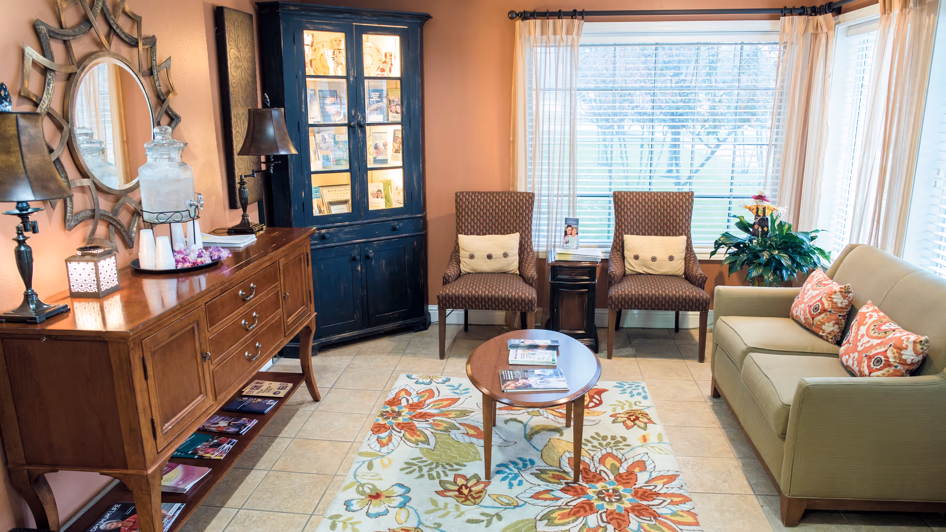 A cozy sitting area with a green sofa adorned with patterned cushions, two patterned armchairs with beige pillows, a wooden coffee table with magazines, a colorful floral rug, a wooden sideboard with lamps and a water dispenser, and a black cabinet with glass doors displaying brochures. Large windows with sheer curtains let in natural light and show a view of trees outside.