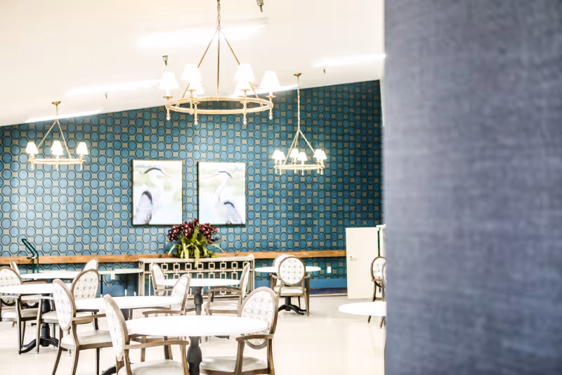 Bright dining room with round tables and patterned chairs, chandeliers, and a teal geometric accent wall featuring bird artwork.