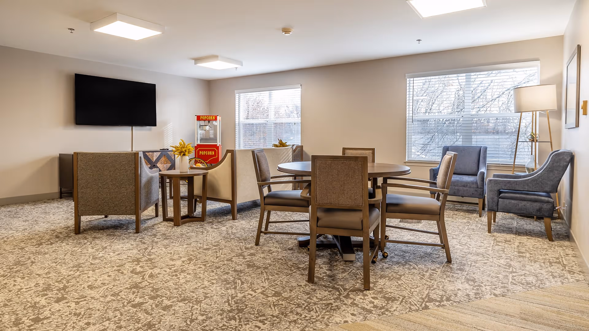 A well-lit common area in a senior living facility with a round table surrounded by four chairs in the center. There are two armchairs and a small table with a vase of yellow flowers near a wall-mounted flat-screen TV. A popcorn machine is visible in the corner near two large windows with blinds. The room has a patterned carpet and a floor lamp beside two additional armchairs near the windows.