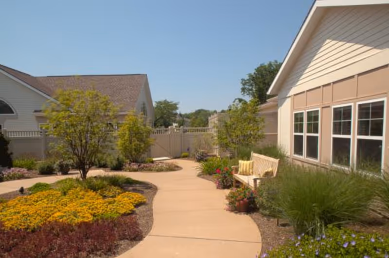 A sunny outdoor garden area at Glenmere Assisted Living featuring a curved concrete pathway, colorful flower beds, small trees, and a wooden bench with a cushion next to a building with multiple windows.