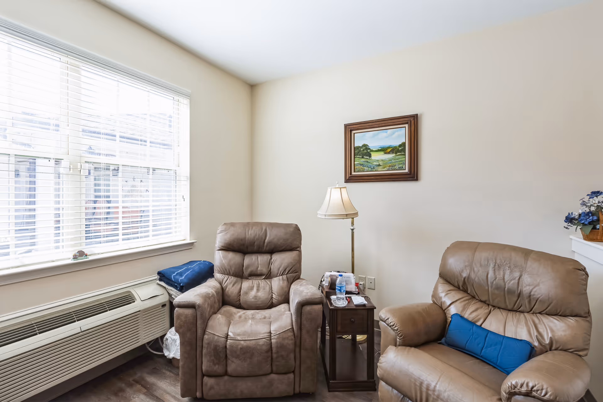 A cozy living room area with two brown leather recliner chairs separated by a small wooden side table holding a lamp, water bottle, and other small items. A window with white blinds allows natural light into the room. A framed landscape painting hangs on the beige wall above the lamp. A blue pillow rests on one of the chairs, and a small plant with blue flowers is on a ledge to the right.