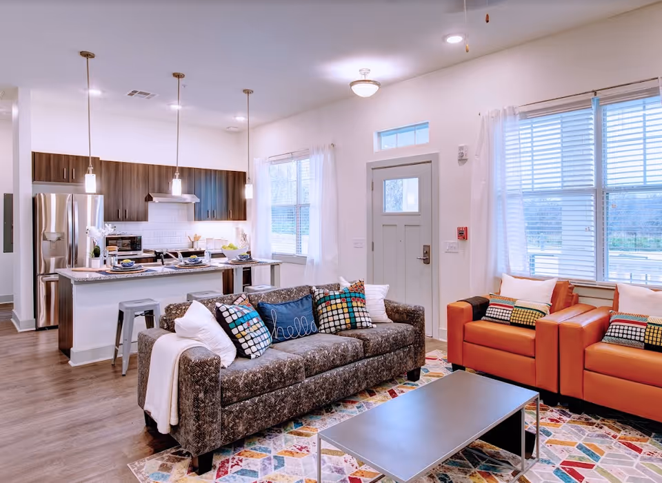 A bright and modern open-concept living room and kitchen area. The living room features a dark patterned sofa with colorful pillows, two orange armchairs with matching pillows, and a rectangular coffee table on a multicolored geometric rug. The kitchen has dark wood cabinets, a stainless steel refrigerator, a white subway tile backsplash, and a granite countertop island with three pendant lights and metal bar stools. Large windows with white curtains allow natural light to fill the space.