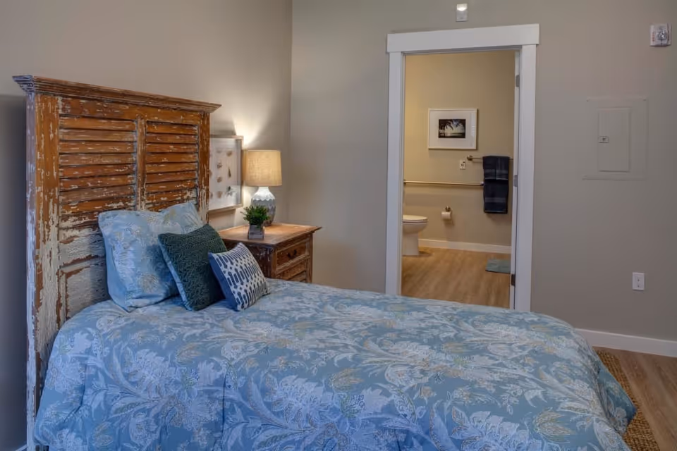 A bedroom with a bed featuring a distressed wooden headboard and blue floral bedding. There are three pillows on the bed in shades of blue and green. A wooden nightstand with a lamp and a small plant is next to the bed. An open door reveals a bathroom with a toilet, a towel rack with a dark towel, and a framed picture on the wall.