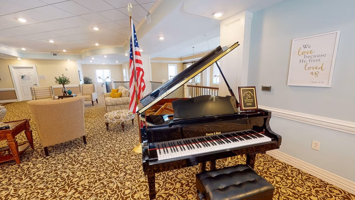 Interior view of a senior living facility lounge area featuring a black grand piano with a bench, an American flag, upholstered chairs, a patterned carpet, and a framed inspirational quote on the wall.