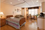 A well-lit senior living facility room featuring a hospital-style bed with a colorful quilt, a wooden nightstand with a lamp, a patterned sofa, a small dining table with two chairs near a window with blinds and a valance, and a kitchenette area with a microwave and coffee maker.