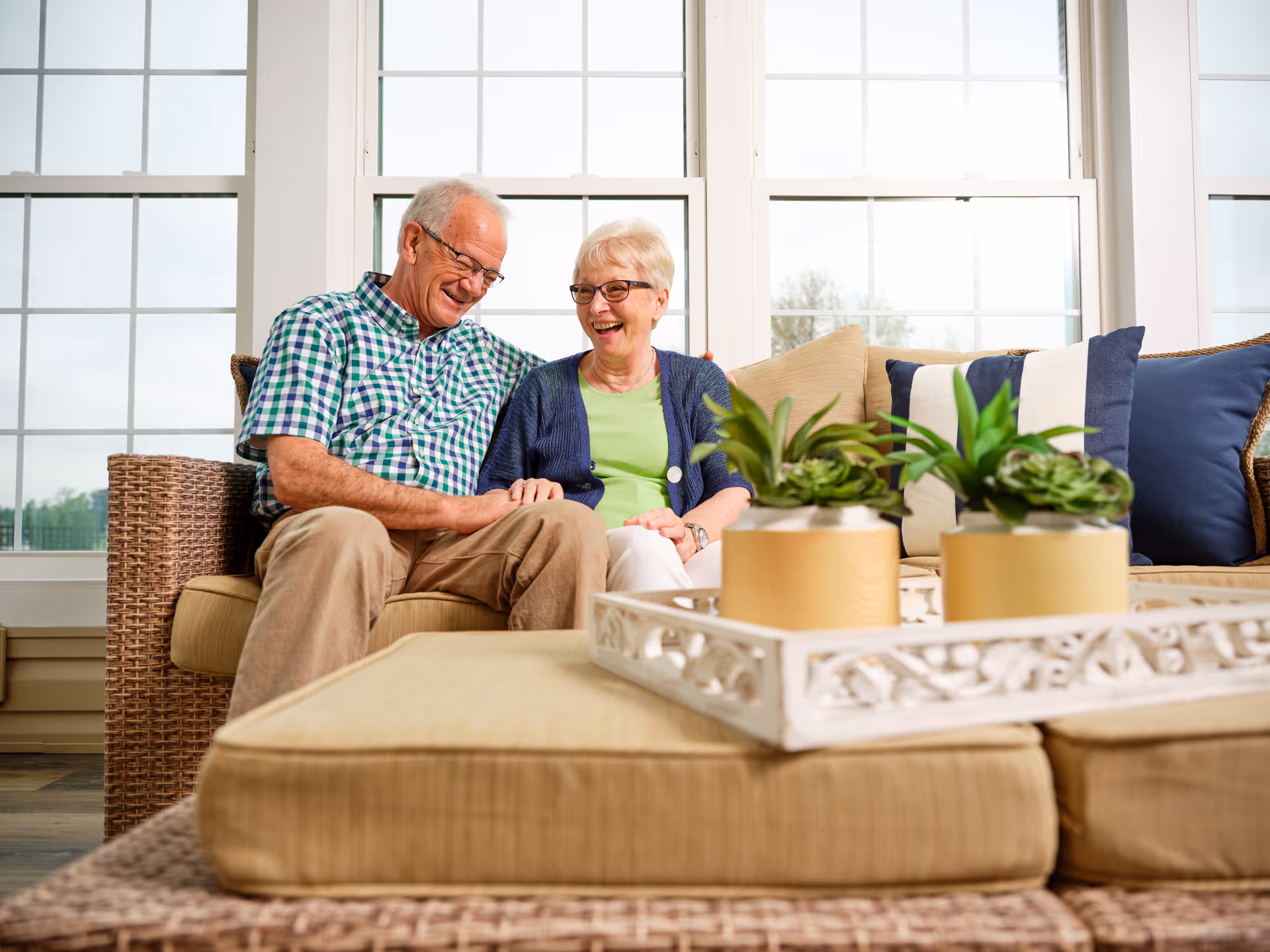 An elderly couple sitting and laughing on a sunlit living room sofa with potted plants on the coffee table.