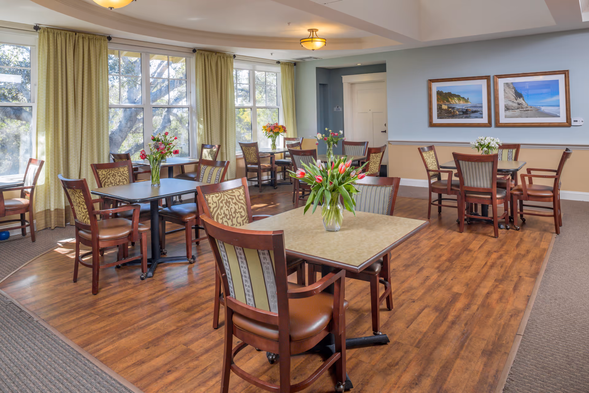 A bright dining room with several square tables and wooden chairs arranged neatly. Each table has a vase with fresh flowers. Large windows with light green curtains allow natural light to fill the room. The walls are painted in soft blue and beige tones, and two framed landscape pictures hang on one wall. The floor is a combination of wood and carpet.