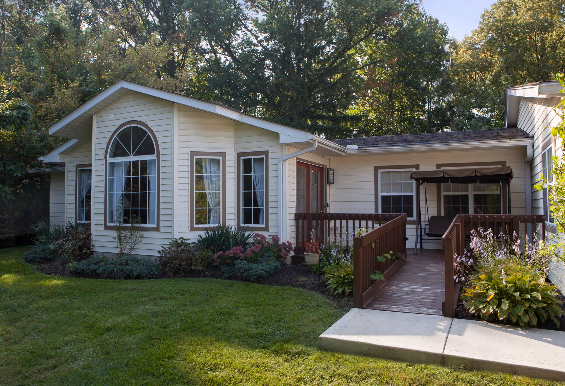 Exterior view of a single-story building with cream-colored siding and brown trim, surrounded by green grass and trees. There is a wooden ramp leading to a porch with a swing seat, and flower beds with various plants and flowers along the building.