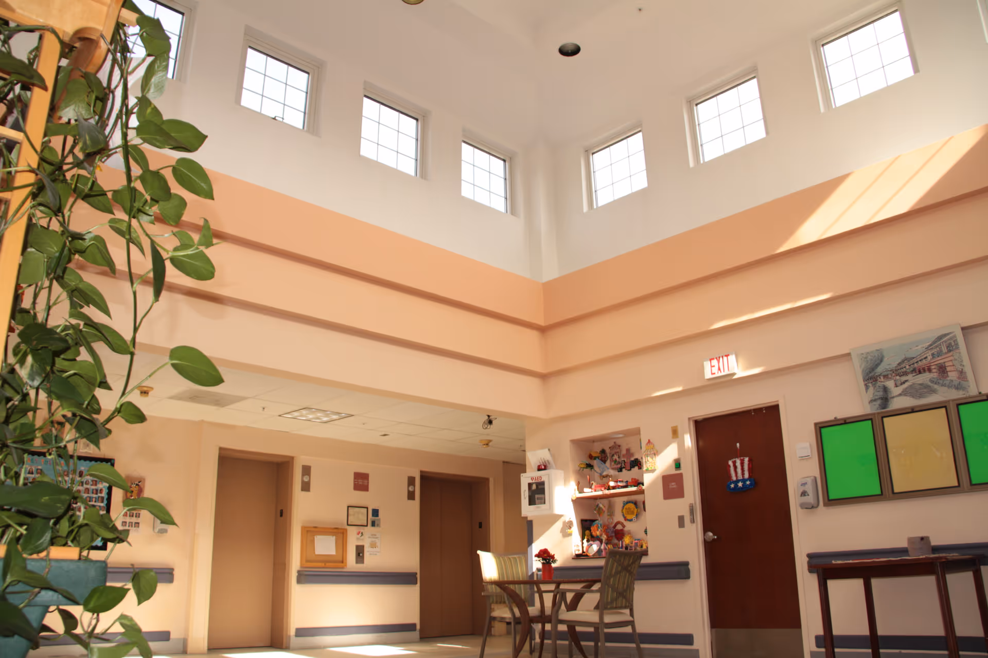 Interior view of a bright common area in a rehabilitation and nursing facility with high ceilings and multiple windows near the ceiling letting in natural light. There are two elevators, a small table with chairs, a plant, and various decorations on the walls including colorful artwork and an exit sign above a door.