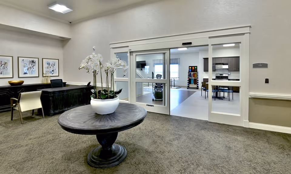 Interior of a senior living common area with a round table and white orchid in the foreground, a reception desk to the left, and a dining/kitchen area visible through sliding glass doors.