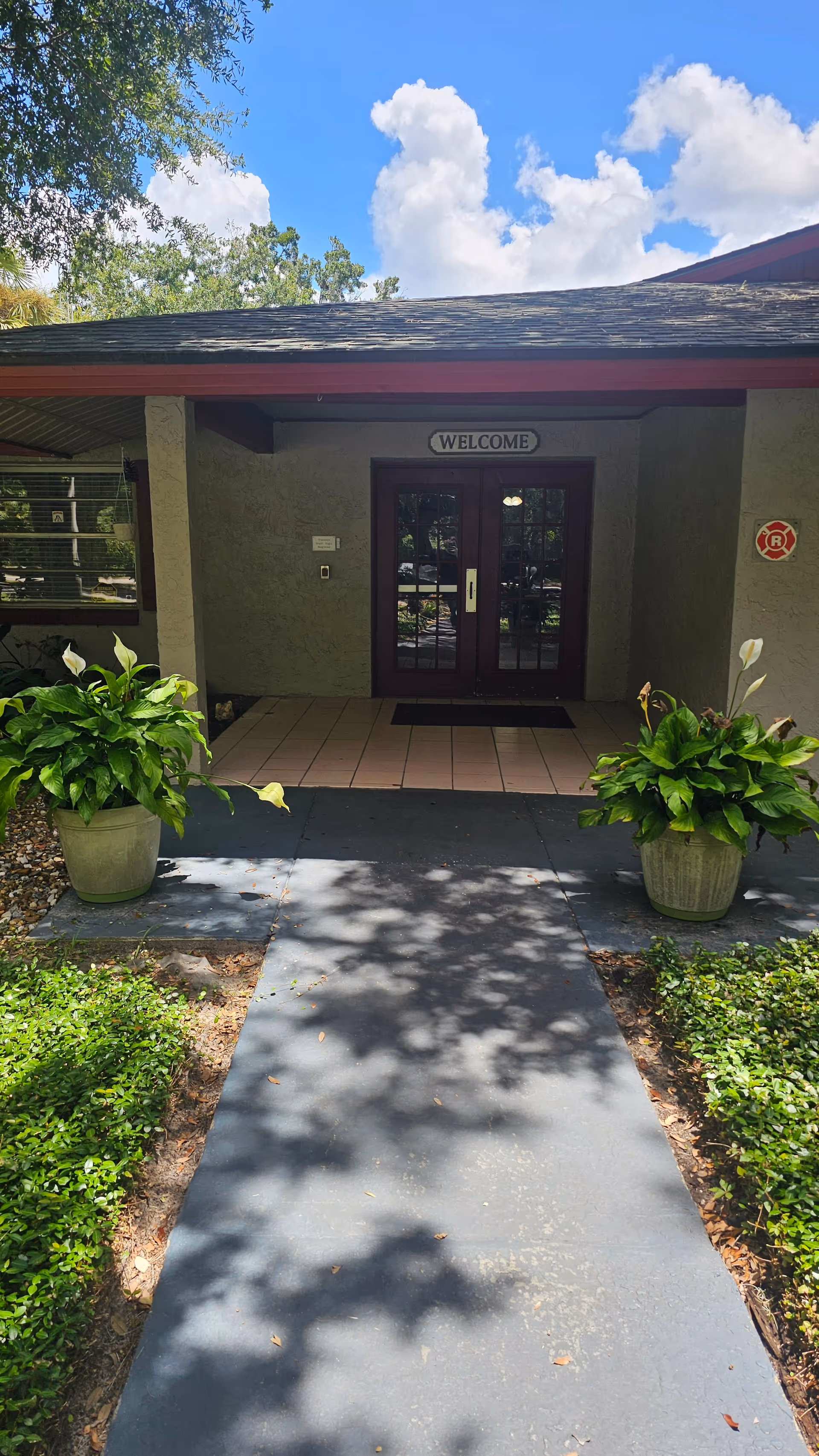 Pathway leading to a covered entrance with double glass doors and a 'WELCOME' sign, flanked by potted plants.