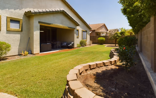 A backyard view of a residential senior living facility showing a well-maintained lawn, a covered patio with outdoor furniture, some bushes, and a stone-bordered garden bed along a brick wall under a clear blue sky.
