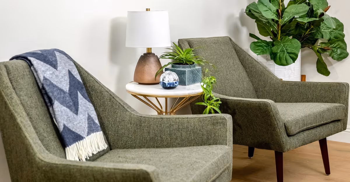 Two green upholstered armchairs with wooden legs placed side by side in a room. One chair has a gray and white chevron patterned throw blanket draped over the back. Between the chairs is a small round side table with a white marble top and gold legs, holding a brown lamp with a white shade, a small green plant in a square pot, and a decorative glass orb. In the background, there is a large leafy green plant in a white pot against a plain white wall.