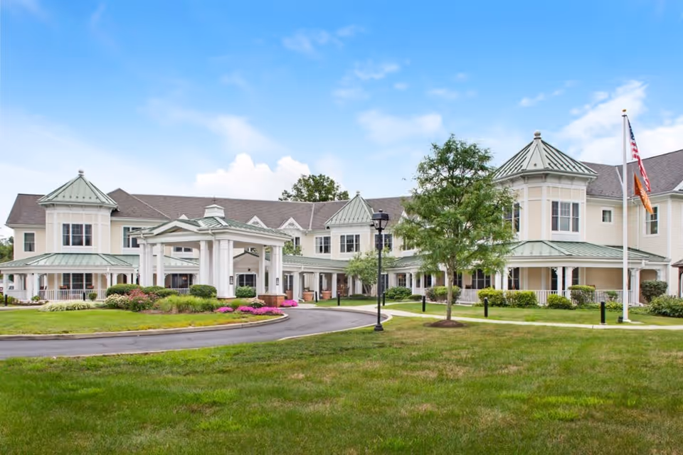 Exterior view of Sunrise of Lower Makefield senior living facility showing a large two-story building with green metal roofs, white columns, and a covered entrance. The building is surrounded by well-maintained lawns, trees, shrubs, and flower beds under a partly cloudy blue sky.