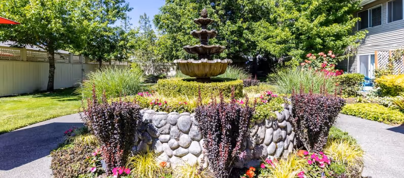 A landscaped outdoor garden area featuring a tiered stone fountain surrounded by various colorful plants and flowers. There is a paved walkway around the garden, green grass, trees, and part of a building visible in the background under a clear blue sky.