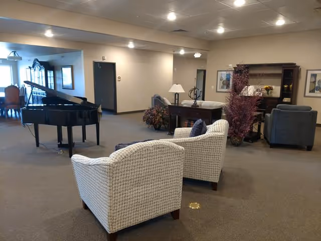 A spacious living room area in an assisted living facility featuring two patterned armchairs in the foreground, a grand piano to the left, a wooden table with a lamp and decorative items, and additional seating including armchairs and a sofa. The room is well-lit with ceiling lights and has neutral-colored walls and carpet.
