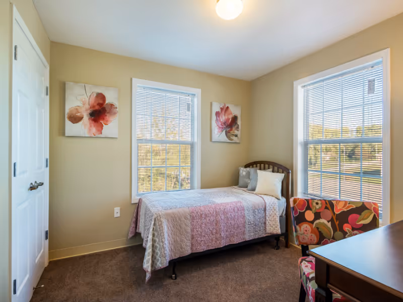 A small bedroom with a single bed covered in a pink and white quilt, two floral paintings on the beige walls, two large windows with blinds letting in natural light, a floral patterned chair, and a wooden desk.