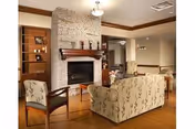 A cozy living room area featuring a stone fireplace with a wooden mantel, a patterned sofa, a wooden chair with floral upholstery, and built-in wooden shelves with decorative items. The room has hardwood flooring and neutral-colored walls with recessed lighting.