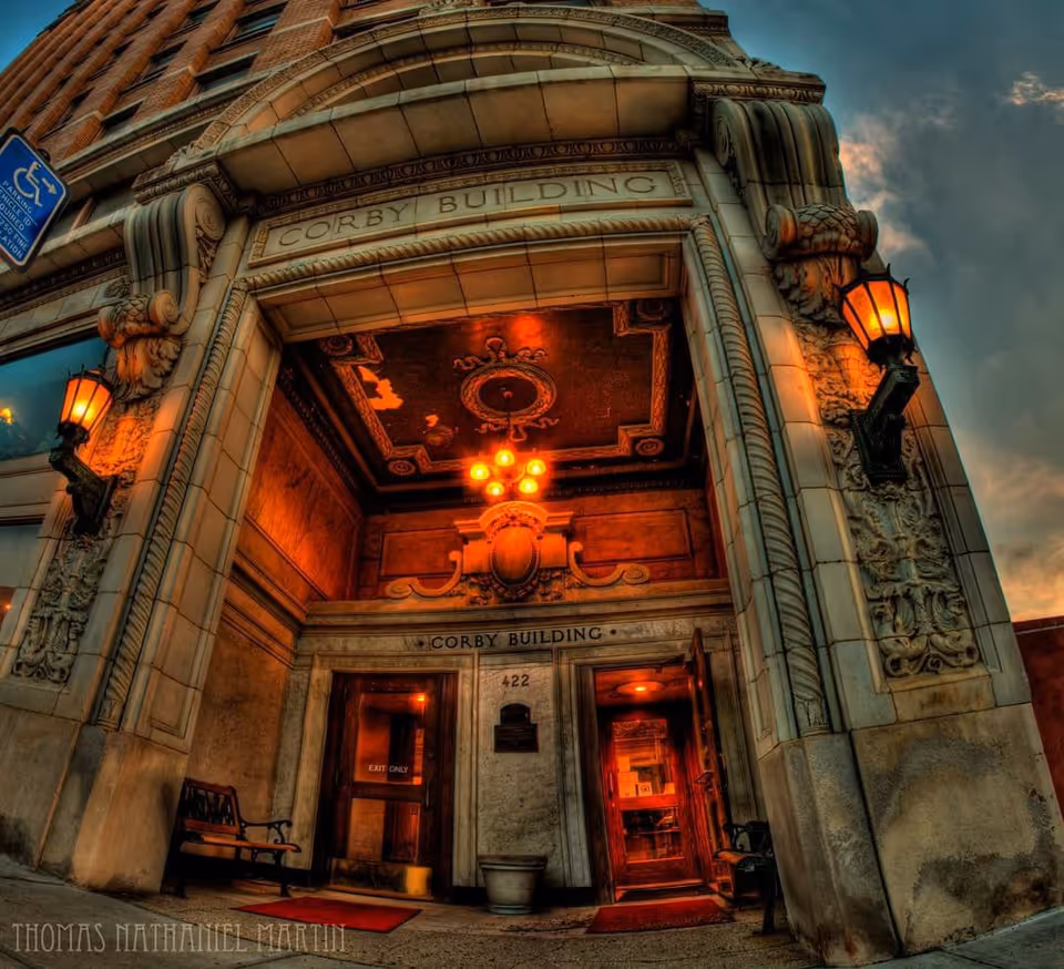Entrance of the Corby Building with ornate architectural details, warm lighting from wall-mounted lanterns, and a decorative ceiling. Two benches are placed near the entrance doors, one labeled 'Exit Only'.