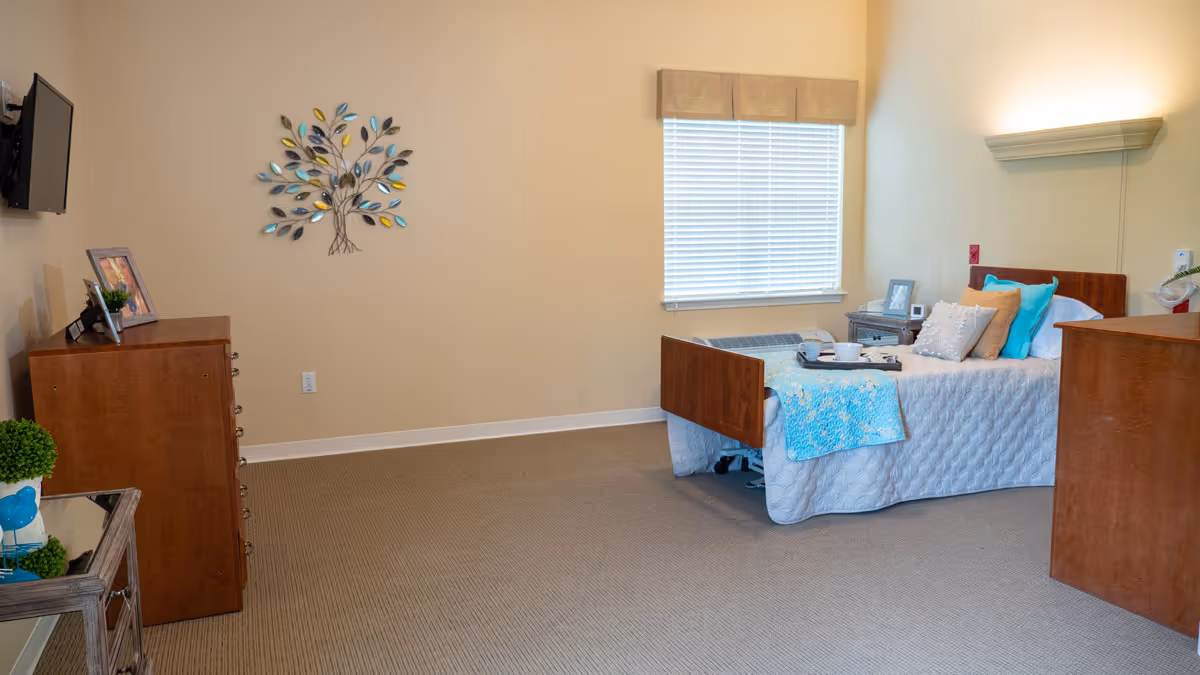 A neatly arranged bedroom in a senior living facility featuring a single bed with white and blue bedding, several pillows, and a tray with cups. The room has beige walls, a window with blinds, a wooden dresser, a small side table with a framed photo, and a wall-mounted TV. Decorative metal wall art shaped like a tree is hung on the wall.