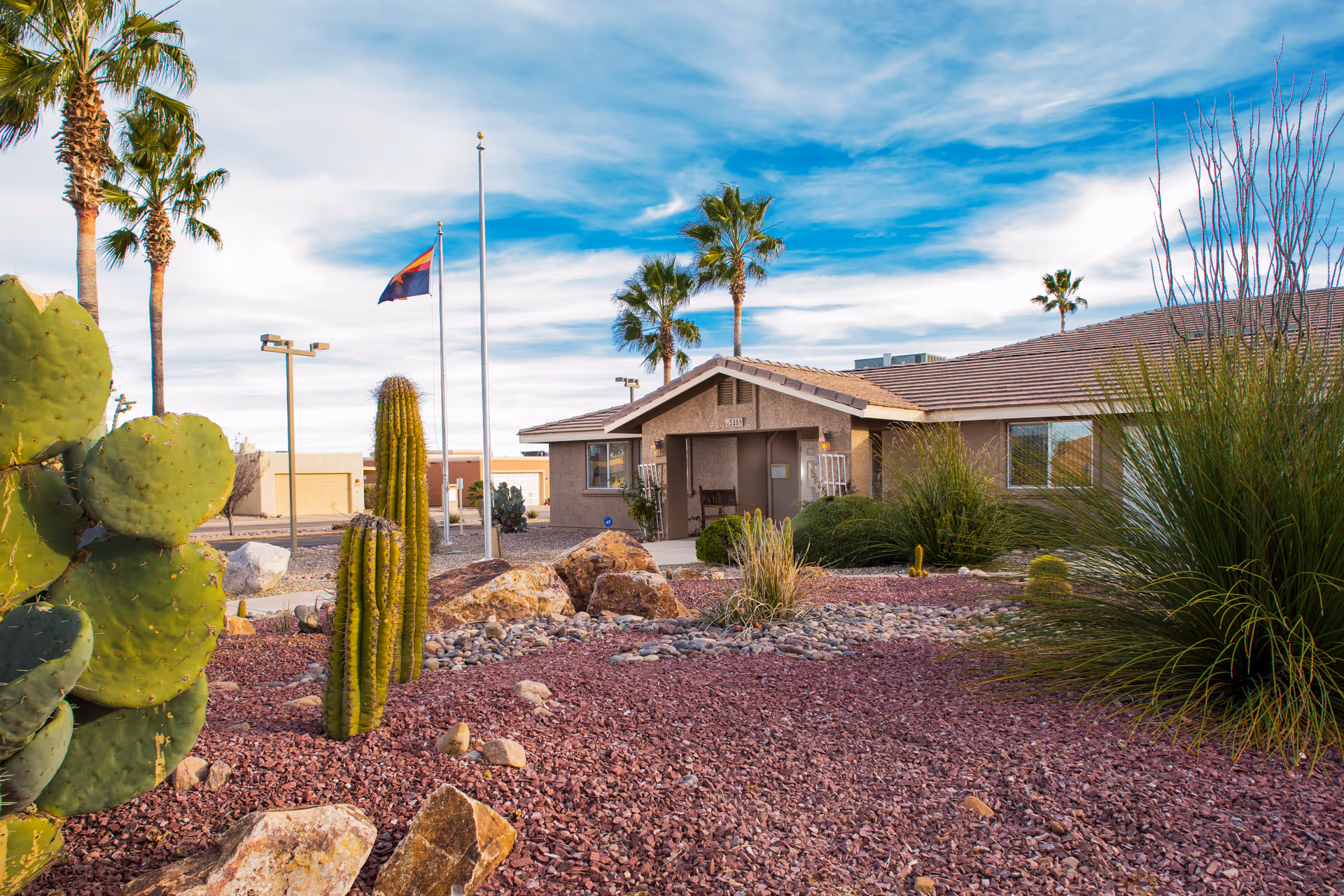 Exterior view of a single-story building with a tiled roof surrounded by desert landscaping including cacti, rocks, and palm trees under a partly cloudy blue sky.