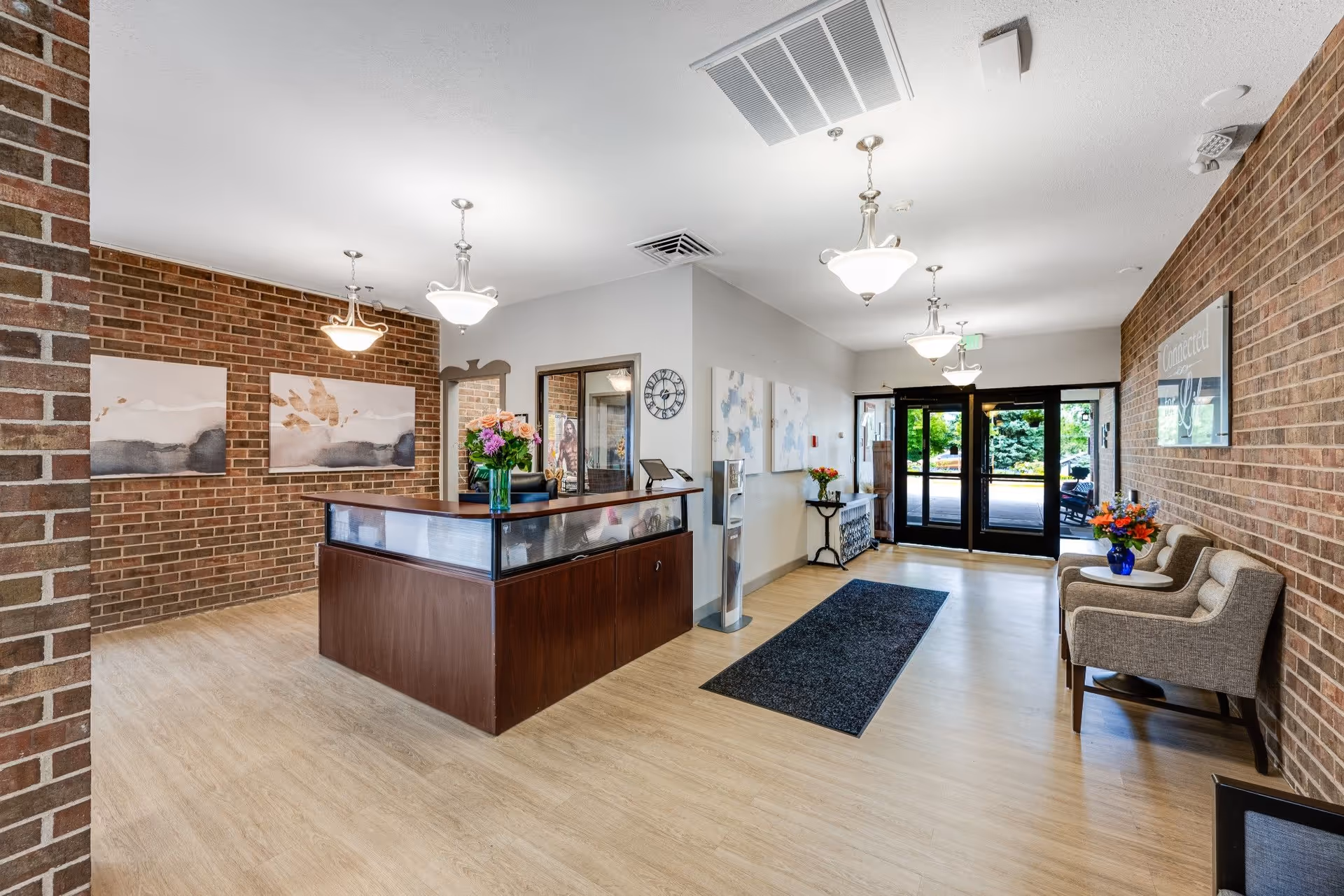 Reception area with a wooden front desk adorned with a vase of flowers, brick walls, modern hanging light fixtures, a clock on the wall, abstract paintings, a water dispenser, and seating area with two armchairs and a small table with a flower vase near the entrance doors.