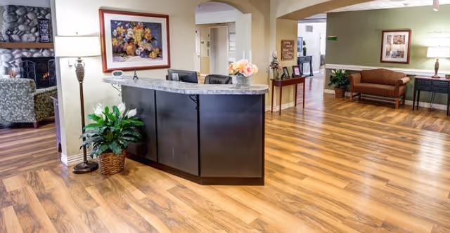 Reception area of a senior living facility with a curved dark reception desk topped with a vase of flowers, a potted plant on the floor, a floor lamp, and framed floral artwork on the wall. The space has wood flooring and an open layout leading to seating areas with chairs and a couch, and a stone fireplace visible in the background.