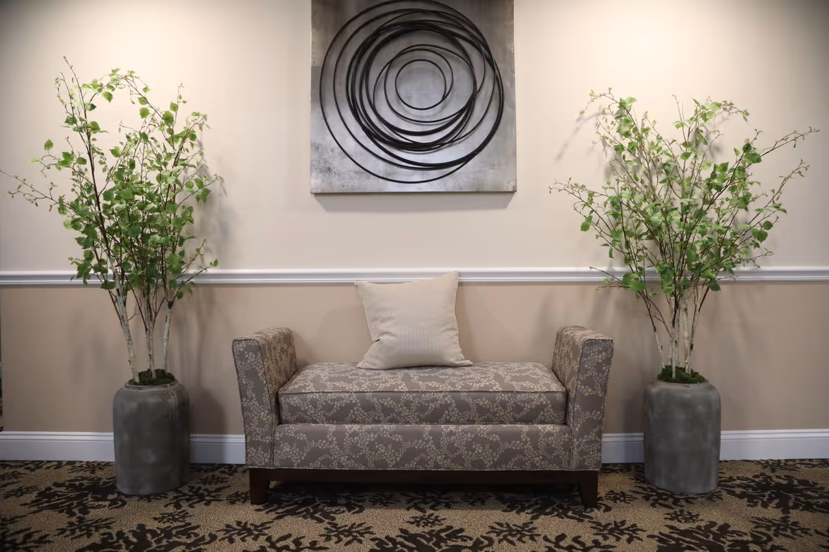 A patterned upholstered bench with a single beige cushion placed in the center, flanked by two large gray vases each holding tall green leafy branches. Above the bench is a modern wall art piece featuring concentric black circular lines on a gray background. The wall behind is painted in two tones with a white chair rail separating the upper and lower sections.